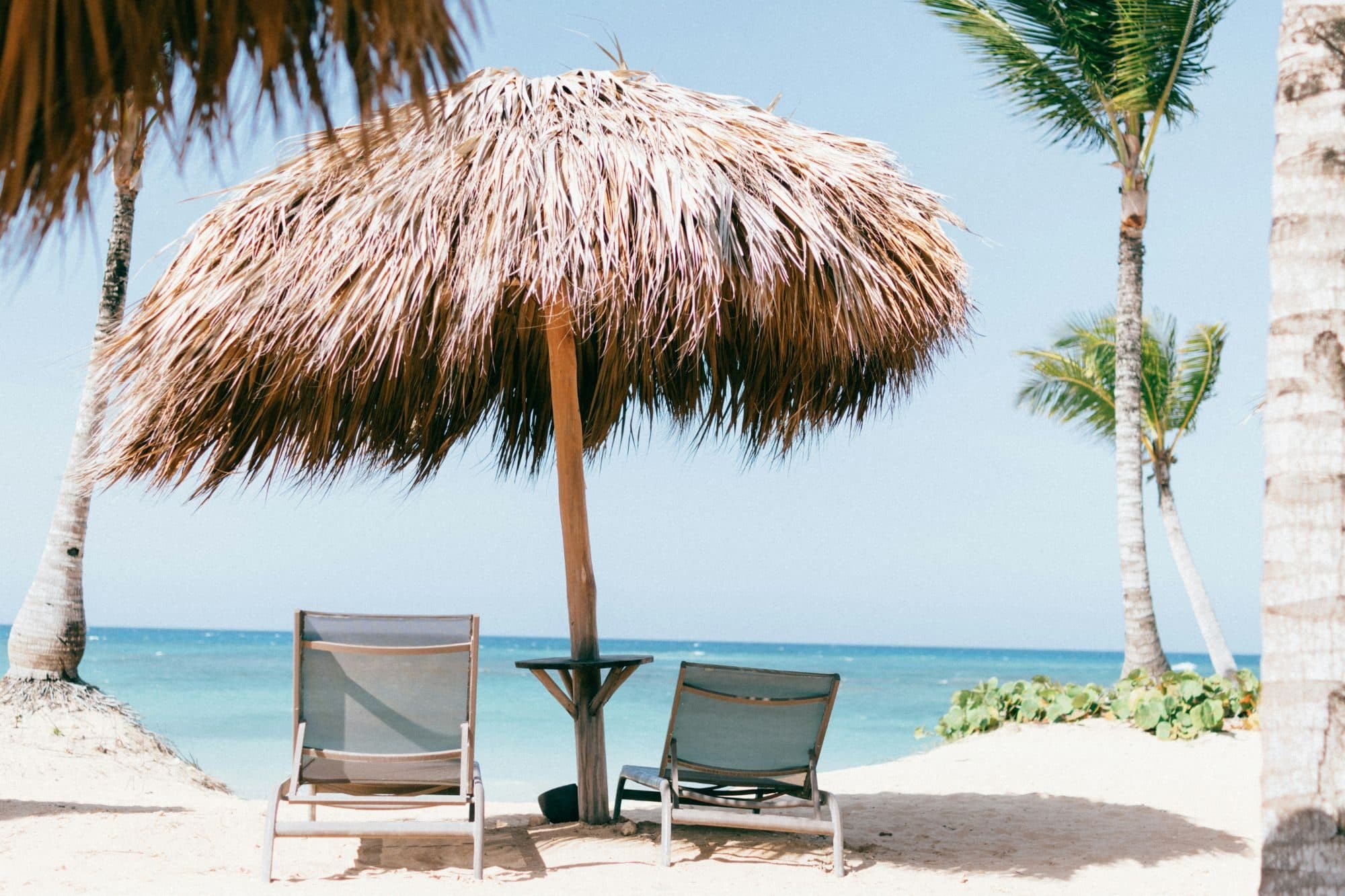 Two sun loungers under a thatched parasol on a white-sand beach by a turquoise sea